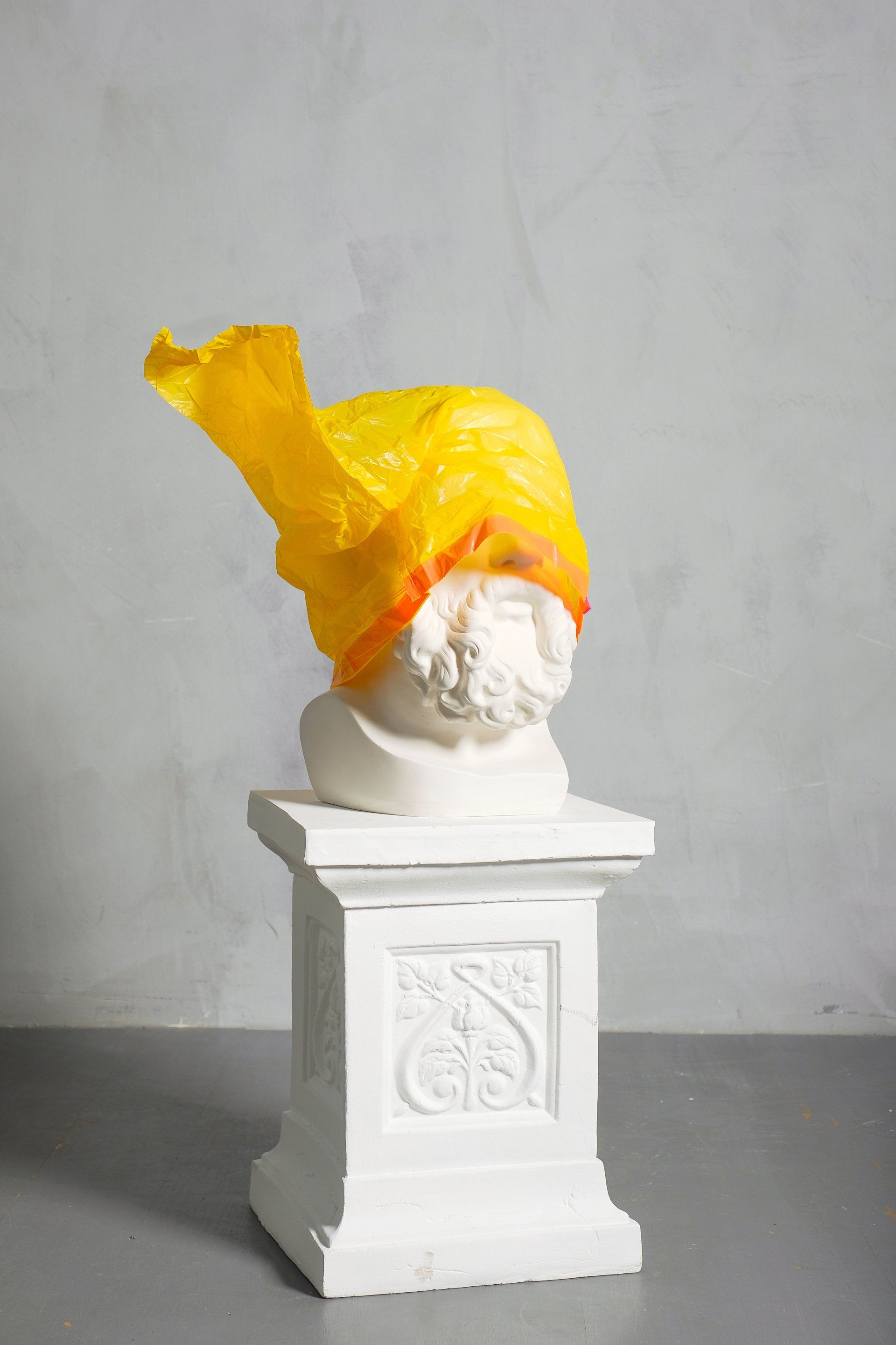 A statue of a man with a yellow bag on his head is sitting on top of a white pedestal.