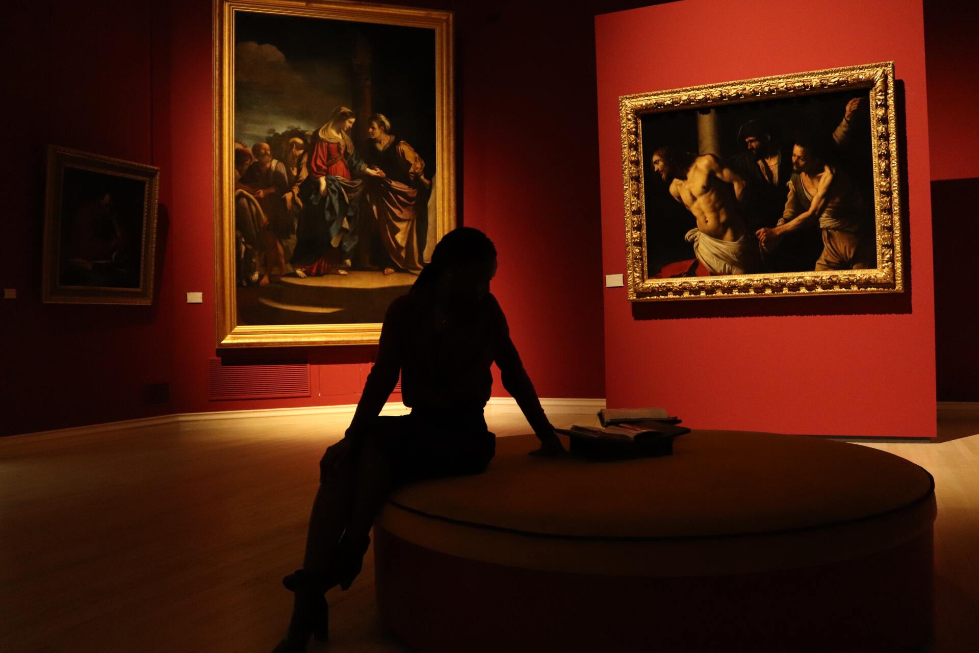 A woman is sitting on a round ottoman in a museum looking at paintings.